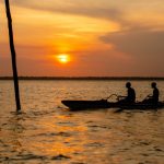 Duas pessoas remam em caiaques no rio ao entardecer, com a paisagem amazônica ao fundo.