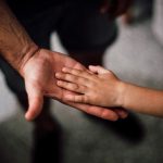 Mão de uma criança repousa sobre a mão de um adulto, simbolizando cuidado, apoio e proteção.
