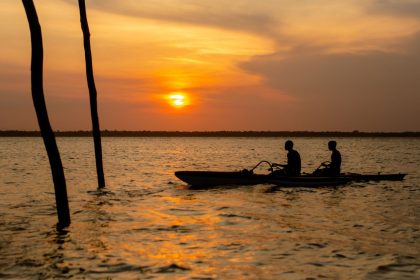 Duas pessoas remam em caiaques no rio ao entardecer, com a paisagem amazônica ao fundo.
